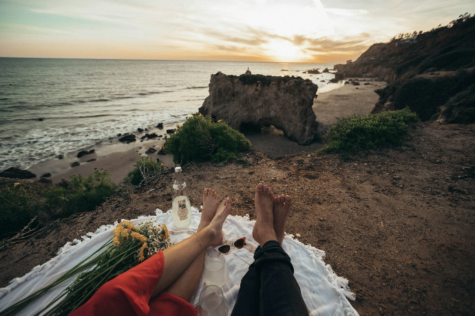 Two people relaxing barefoot on a white blanket overlooking the ocean at sunset